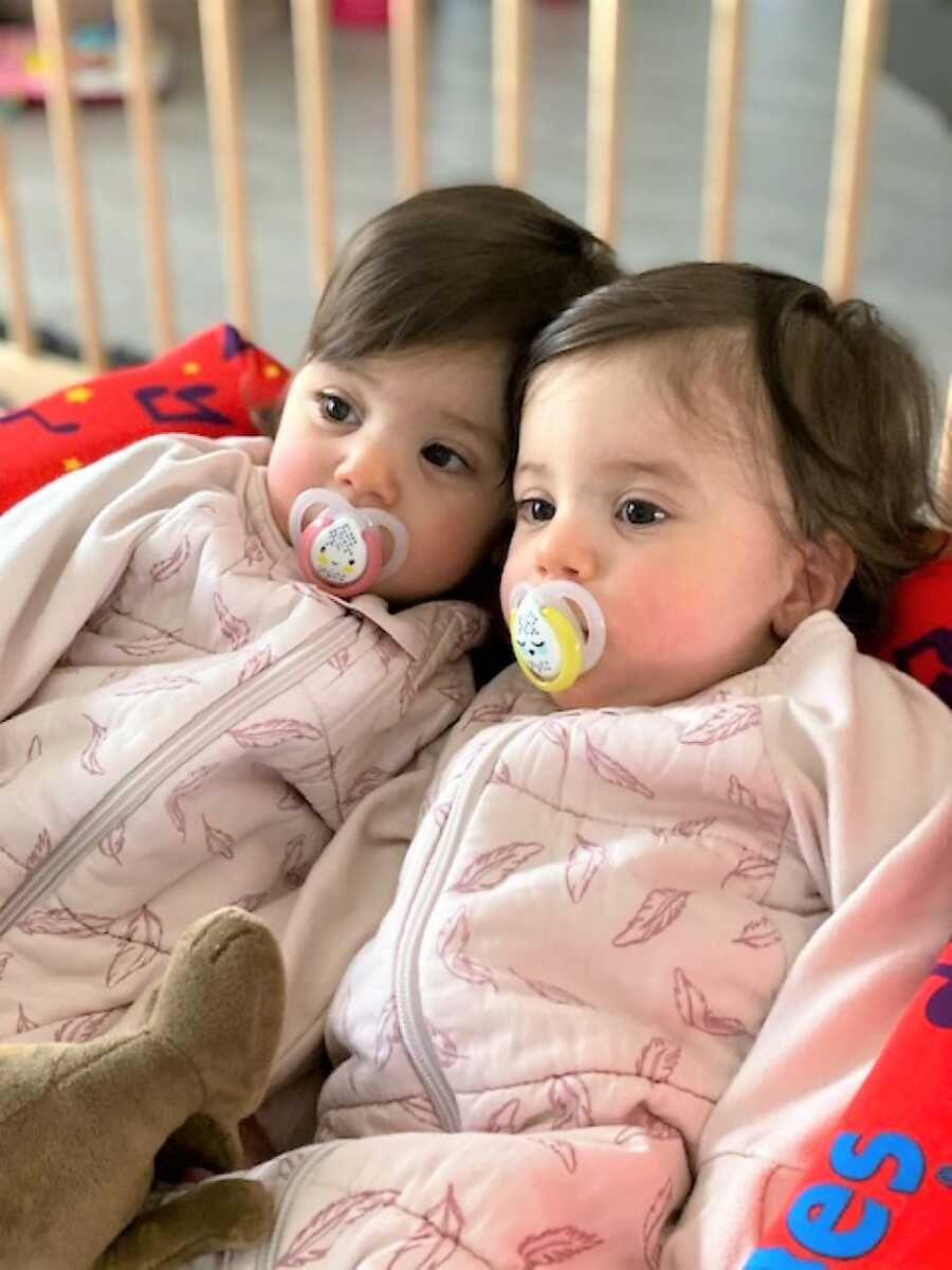 Twin sisters wearing pink matching outfits with pacifiers in their mouths 