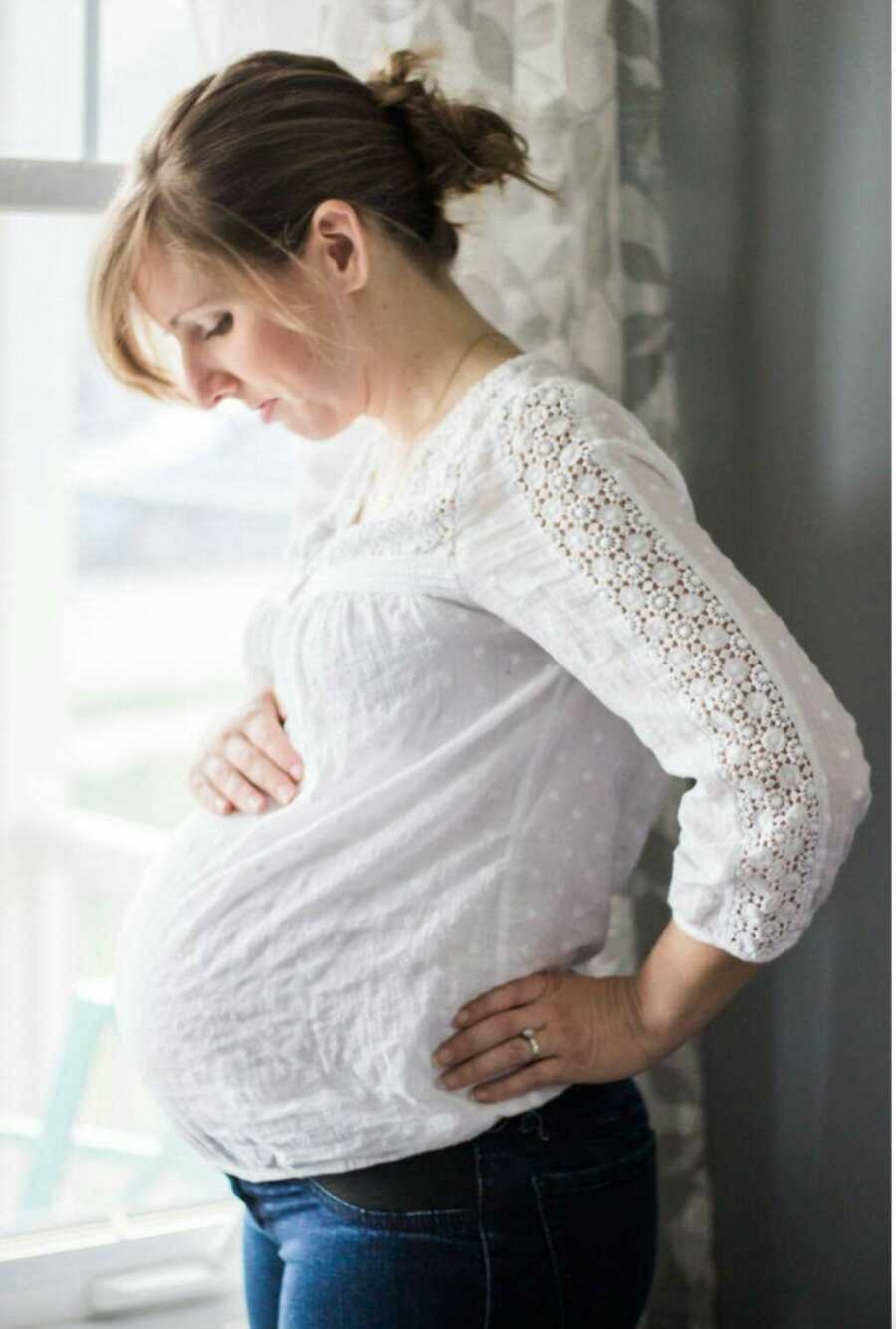 pregnant woman holds and looks down at belly while standing in front of a window 