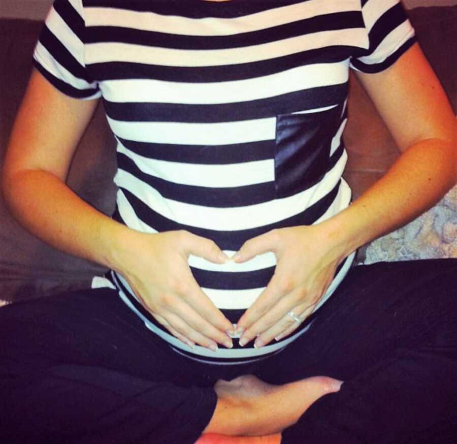 pregnant woman holds hand in heart-shape on top of her pregnant belly while wearing black and white tripped shirt 