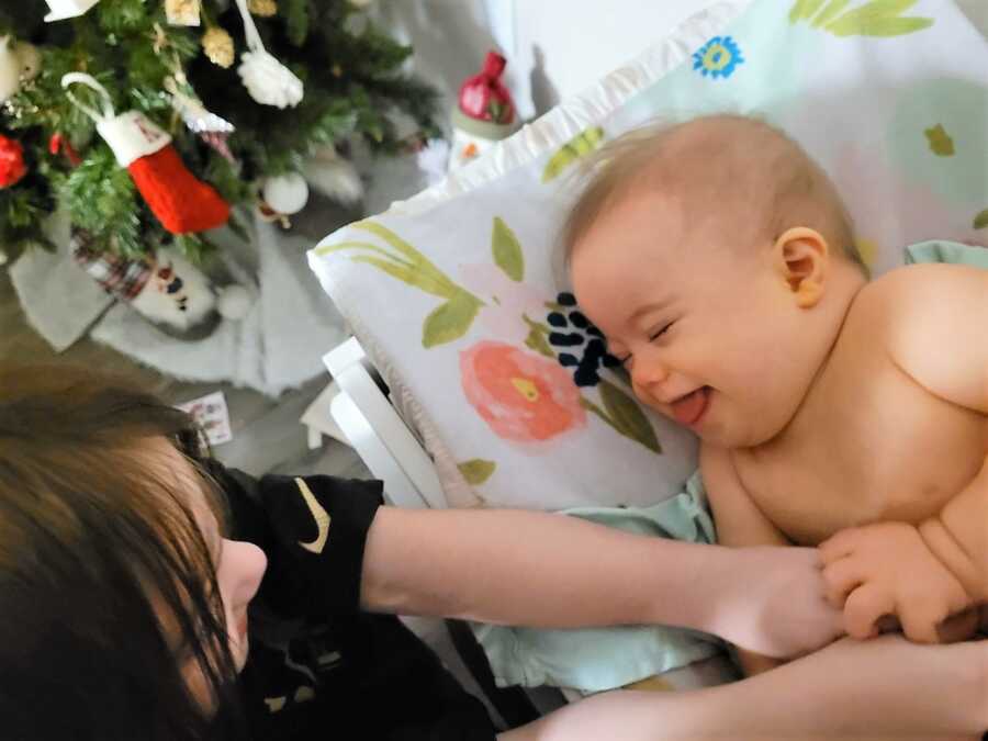 big sister tickling baby sister with down syndrome next to Christmas tree 