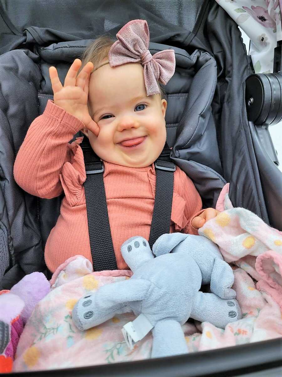 Rainbow baby girl with Down Syndrome sitting on a stroller with a stuffed elephant on her lap and a big pink bow on her head