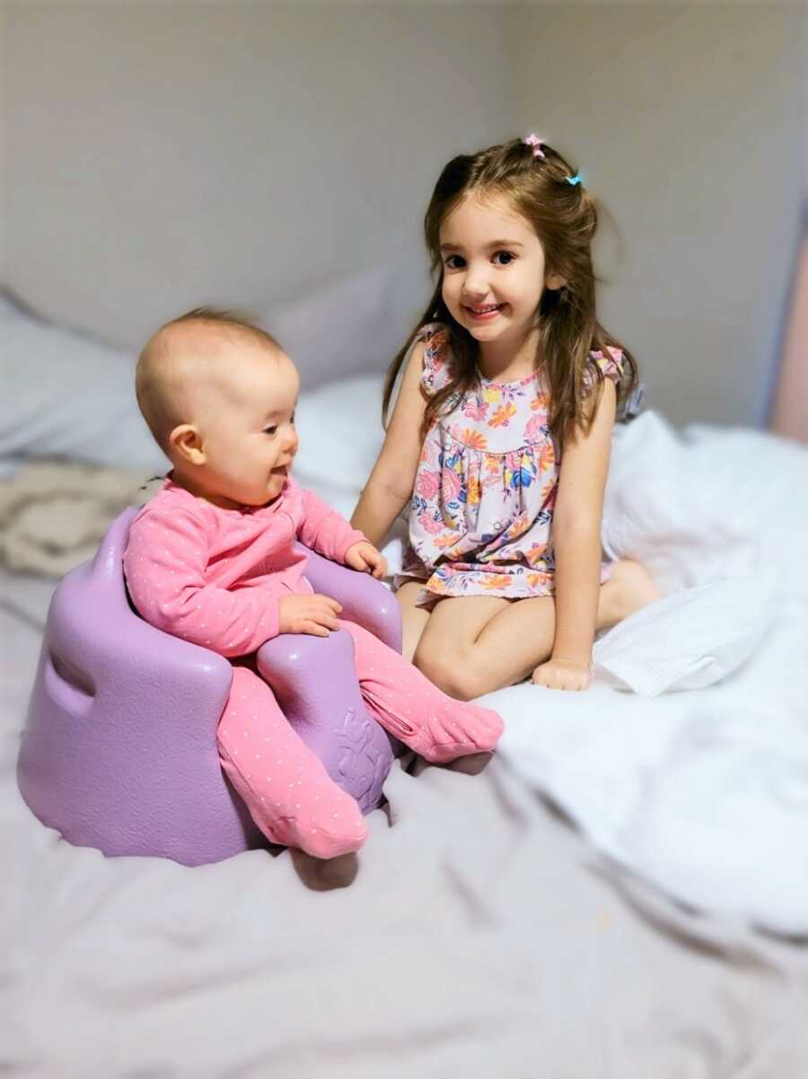 Big sister sitting next to baby sister with Down Syndrome sitting on a purple infant chair 