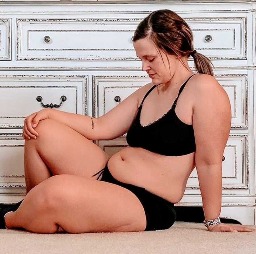 Mom looks down at her postpartum body while wearing black undergarments