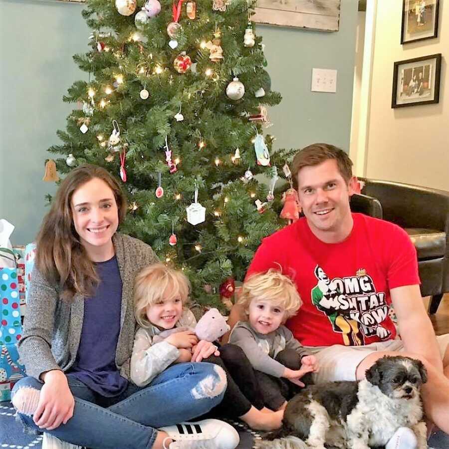 married couple sitting in front of the Christmas tree with their 2 children and the dog