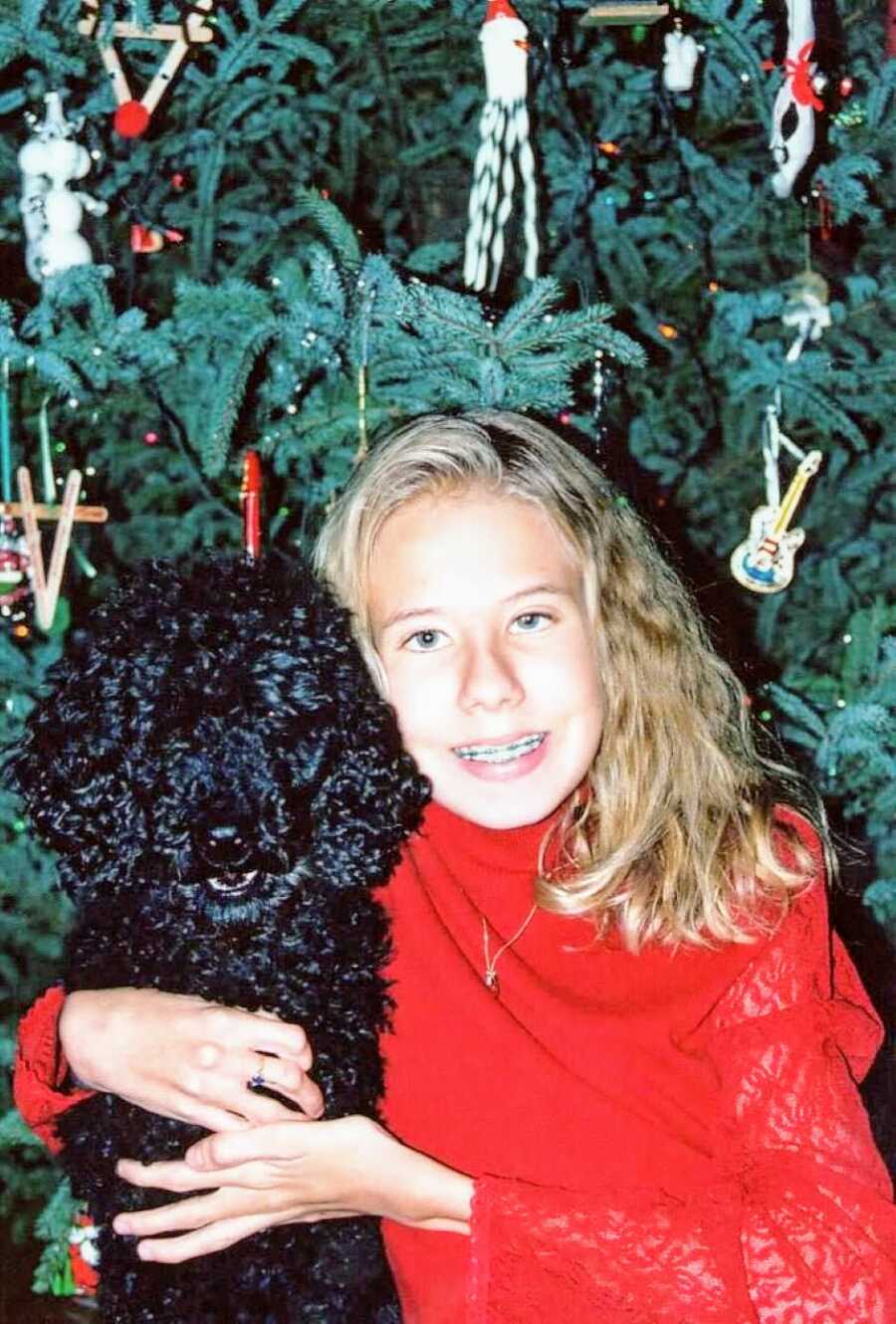 Young girl poses in front of the Christmas tree in a red sweater while hugging a black poodle