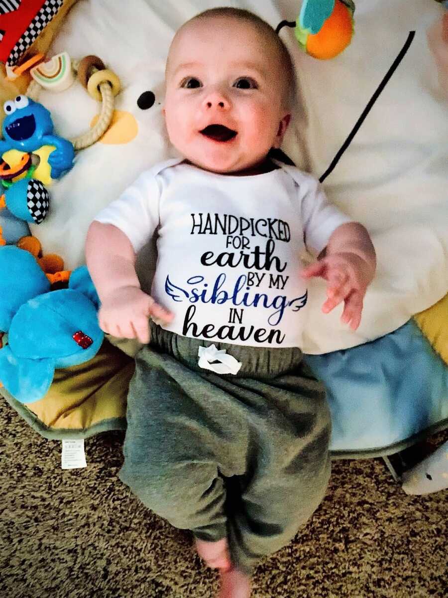 Rainbow baby smiling and wearing a onesie that says HANDPICKED FOR EARTH BY MY SIBLING IN HEAVEN