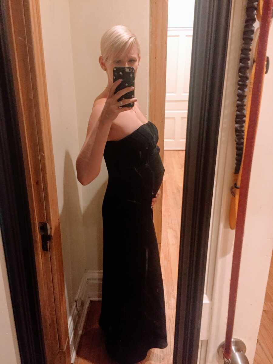 Woman with short blonde hair takes a mirror selfie in a long black sleeveless dress, showing off her belly bump