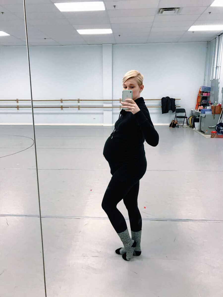 Dance instructor takes selfie of her belly bump in dance studio wearing all black outfit