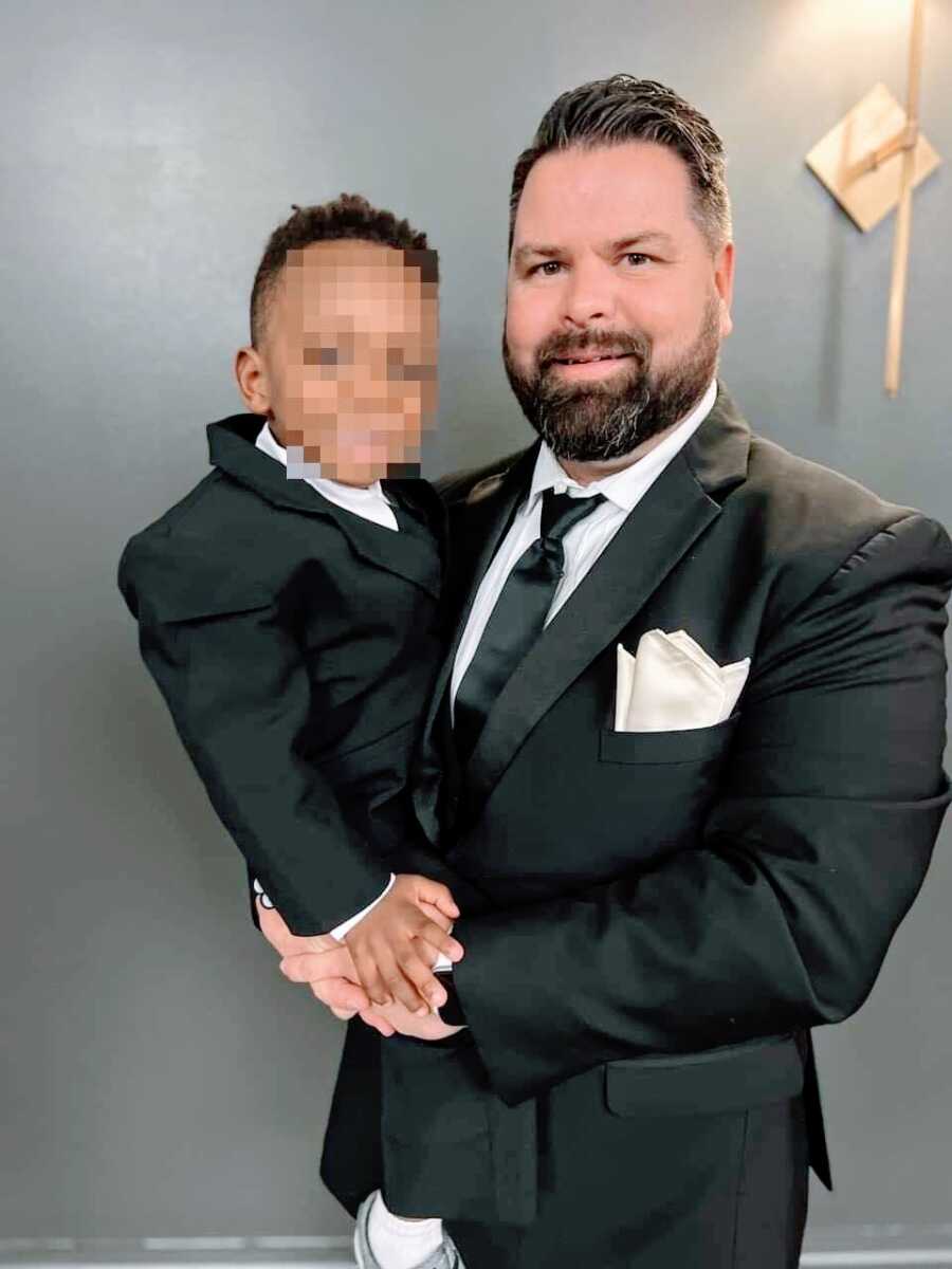 Dad holds foster son while both wearing black suits at fancy Christmas event