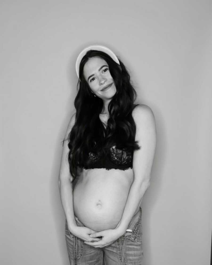 Woman in bra and jeans smiles big while holding her pregnancy belly