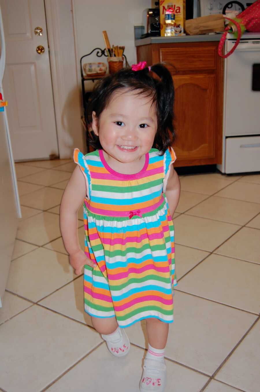 Small Chinese girl in colorful striped t-shirt dress stands in kitchen.