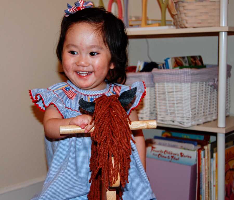 Chinese toddler wears blue dress with American flags and rides rocking horse.