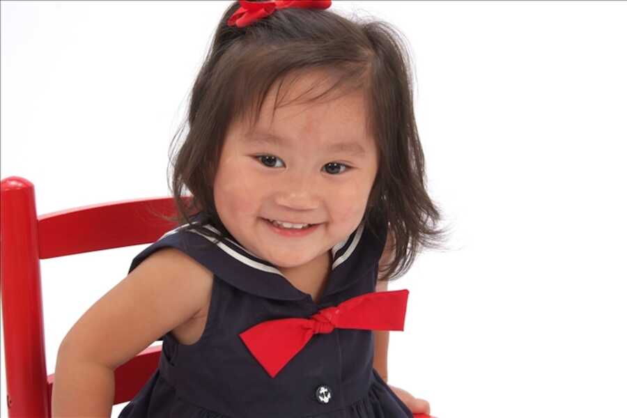 Little Chinese girl wearing a navy sailor's dress sits in red chair for picture.