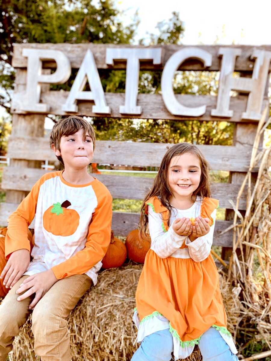 siblings sit on hay bails in festive fall outfits holding pumpkins