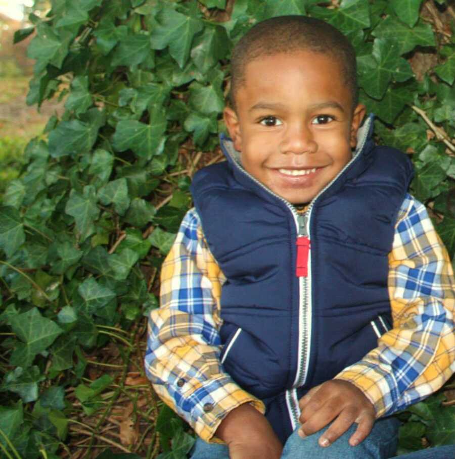 Young boy of single mom wears plaid shirt and jacket vest while smiling.