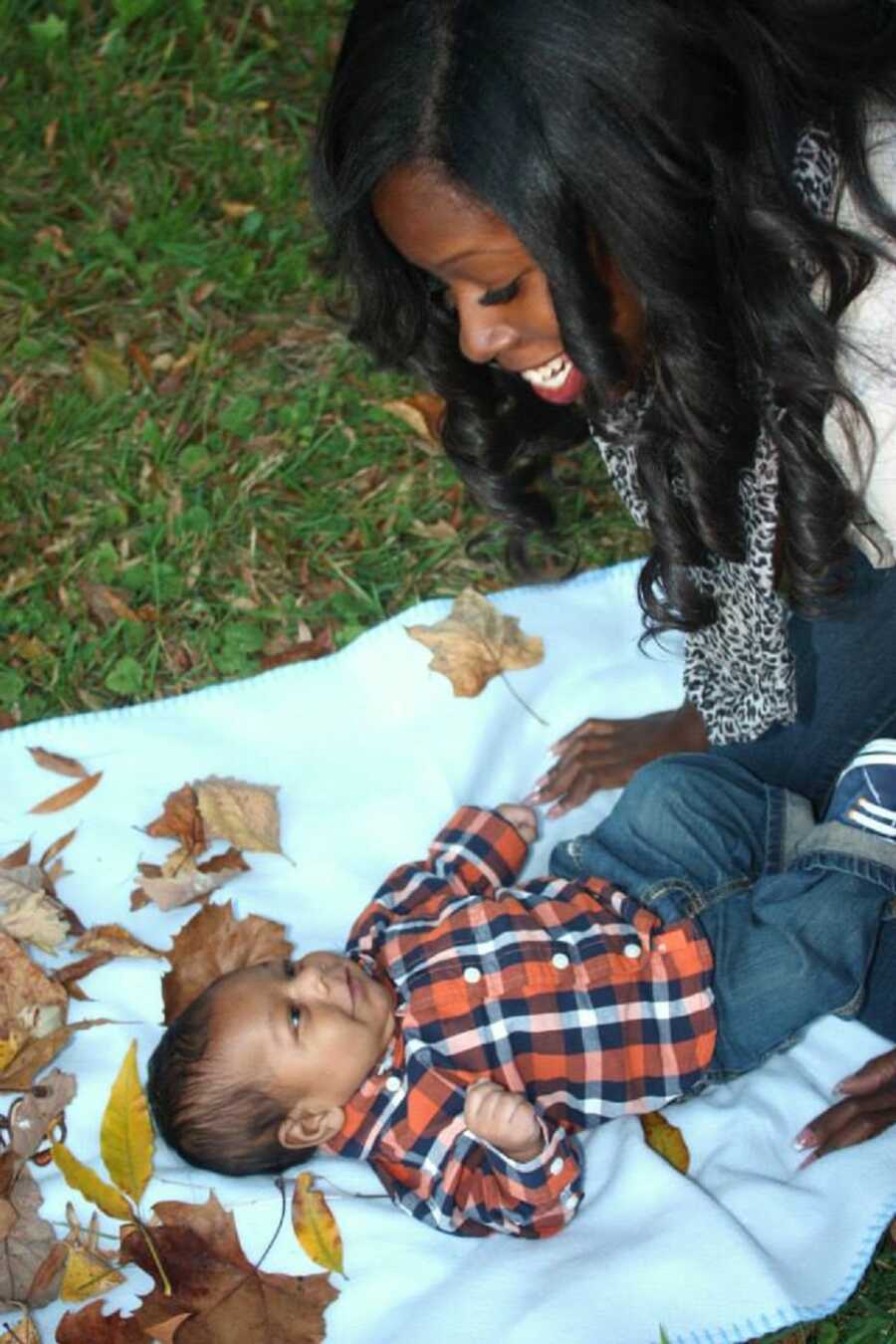 Single mom smiles over toddler boy laying on blanket in the park.