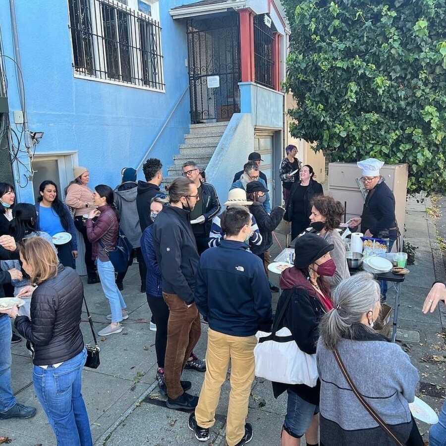 Crowd of people stand outside, eating pancakes and visiting with each other. 