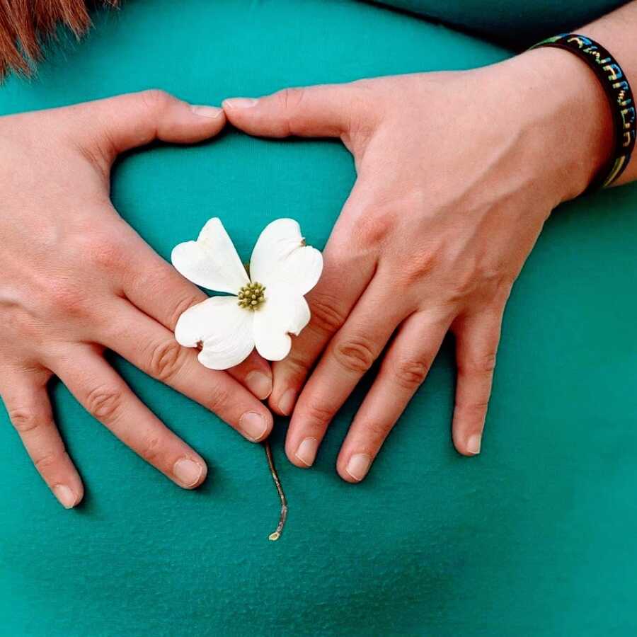 A mom's pregnant belly in a blue dress with hands and a white flower held over it