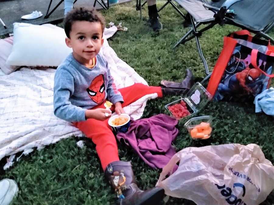 A little boy sits on a blanket on the grass surrounded by food and chairs