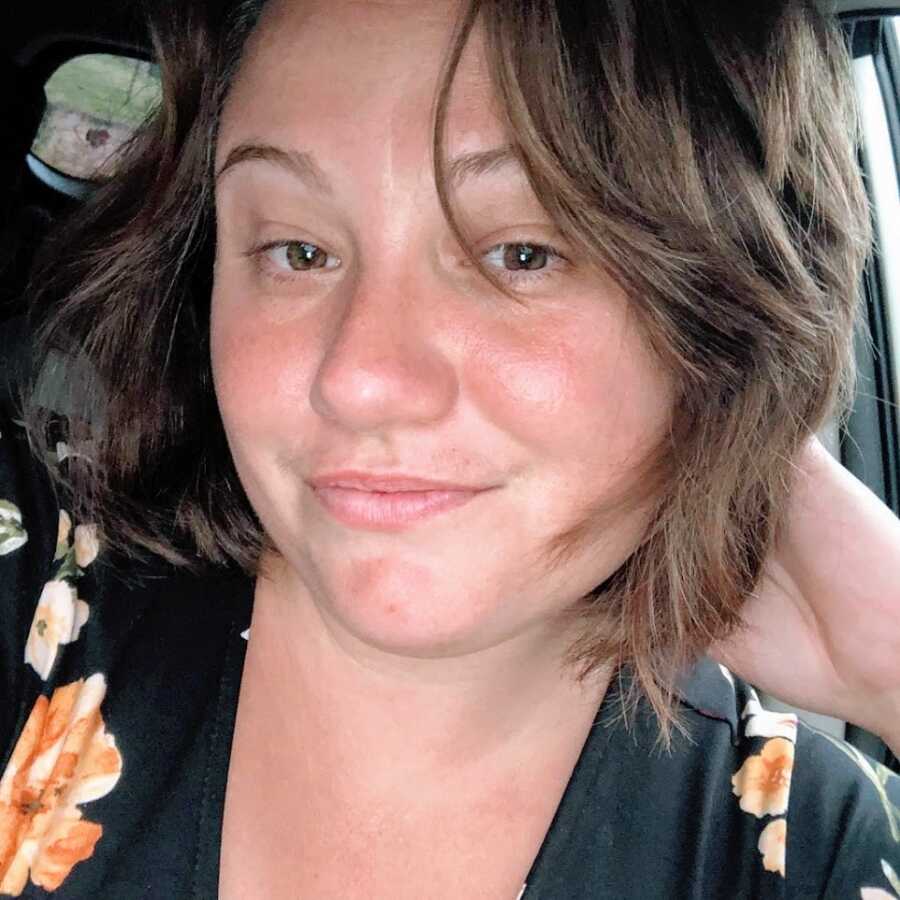 A brown-haired single mom in her car wearing a black shirt with orange flowers