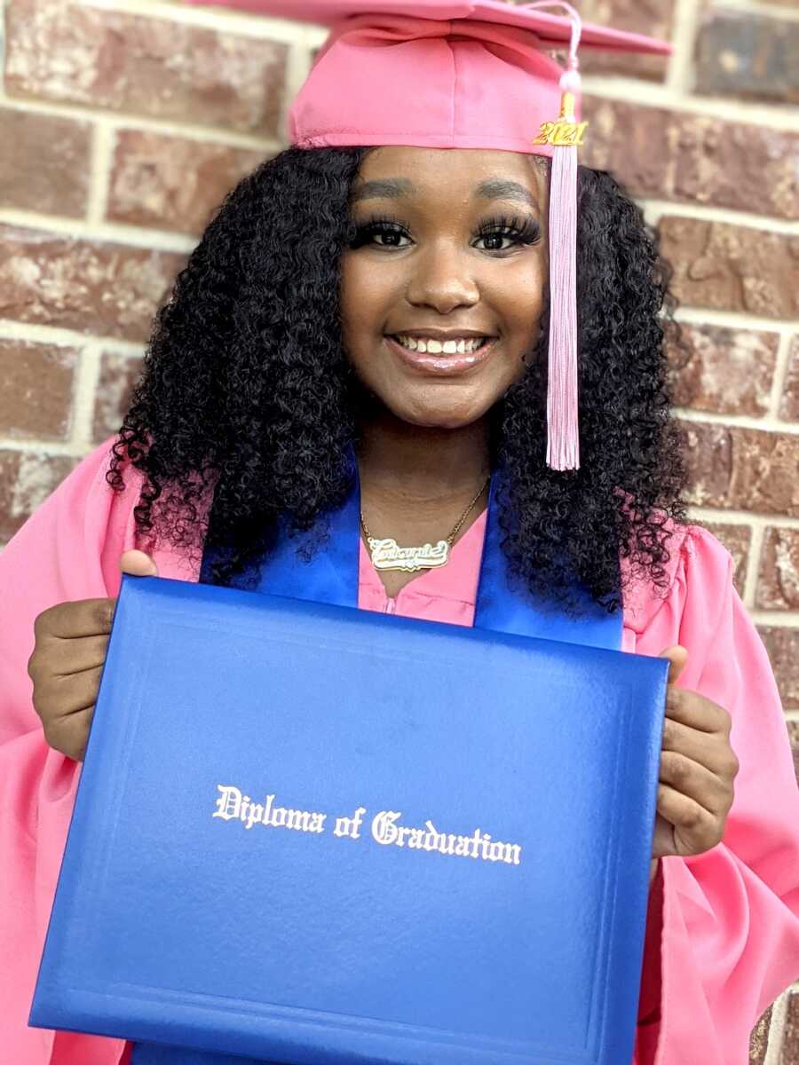daughter of teen mom poses with her high school graduation diploma in a cap and gown