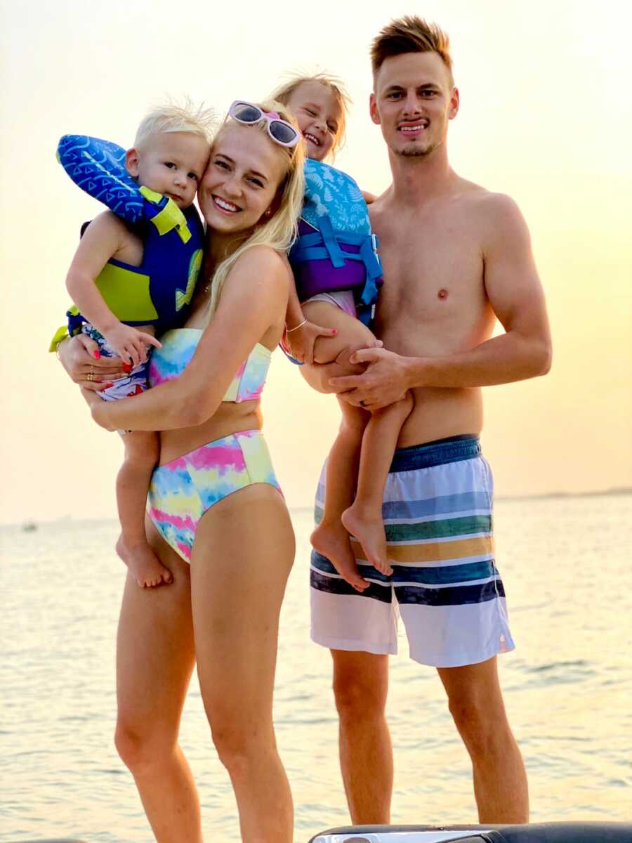 mom and dad hold their son and daughter in their arms while at the beach in swimsuits
