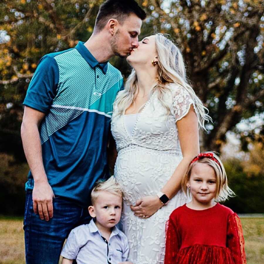 Pregnant woman and husband kiss in front of their two children for family picture.