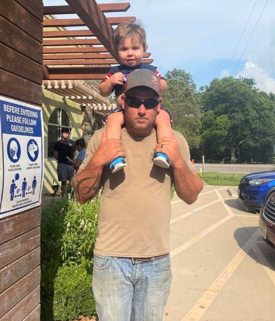 Dad who volunteers as a local firefighter holds toddler son on his shoulders.