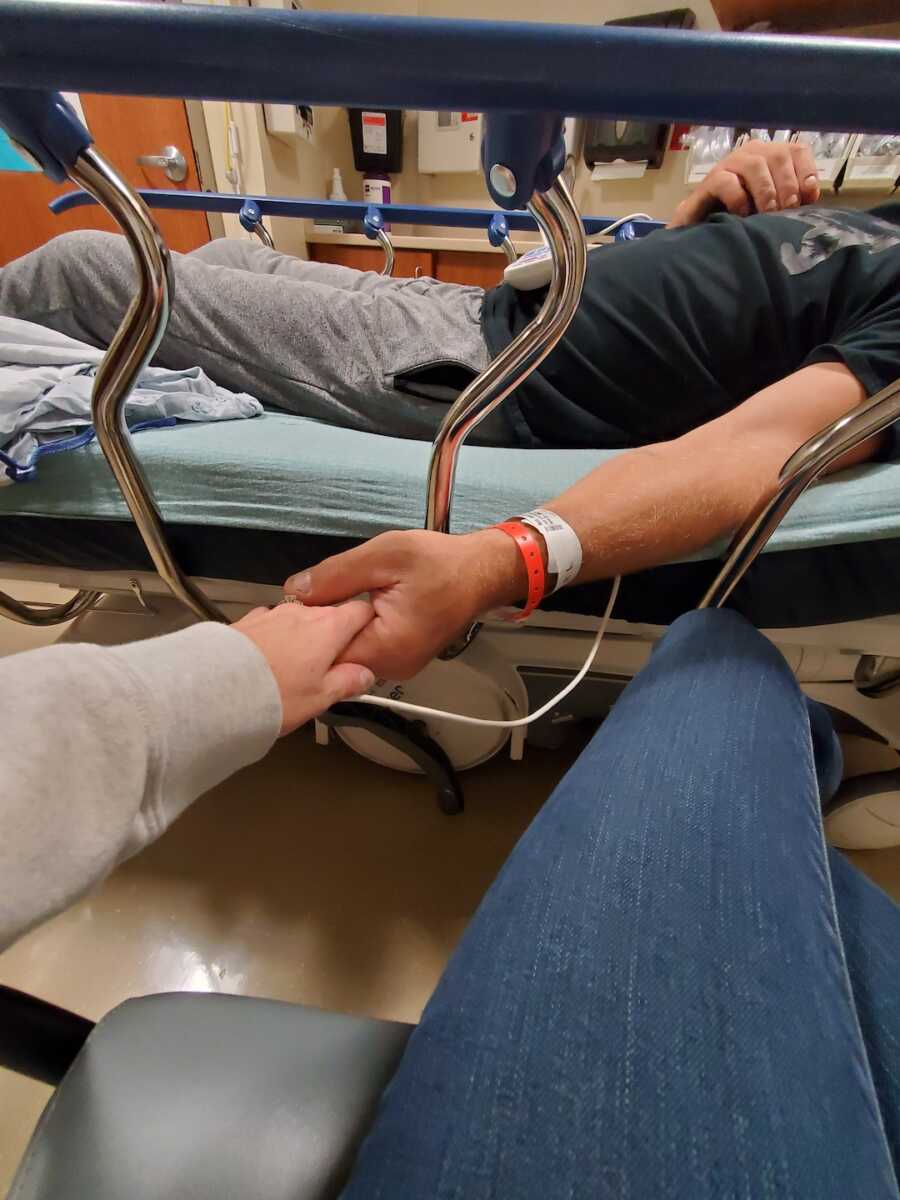 wife holds the hand of her husband while he sits on a gurney in the hospital