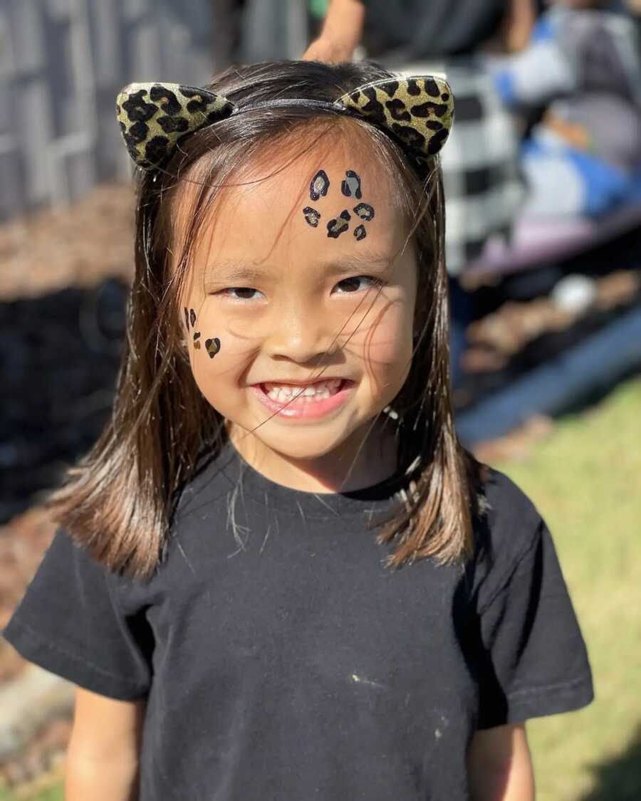 Little girl adopted from Chinese orphanage wears cheetah face paint and cat ears.