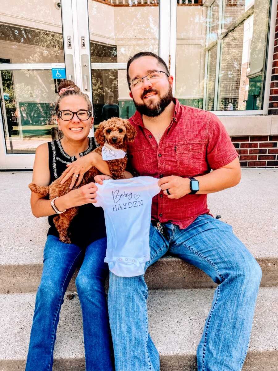 Couple take photos with their dog while holding a onesie announcing their adoption