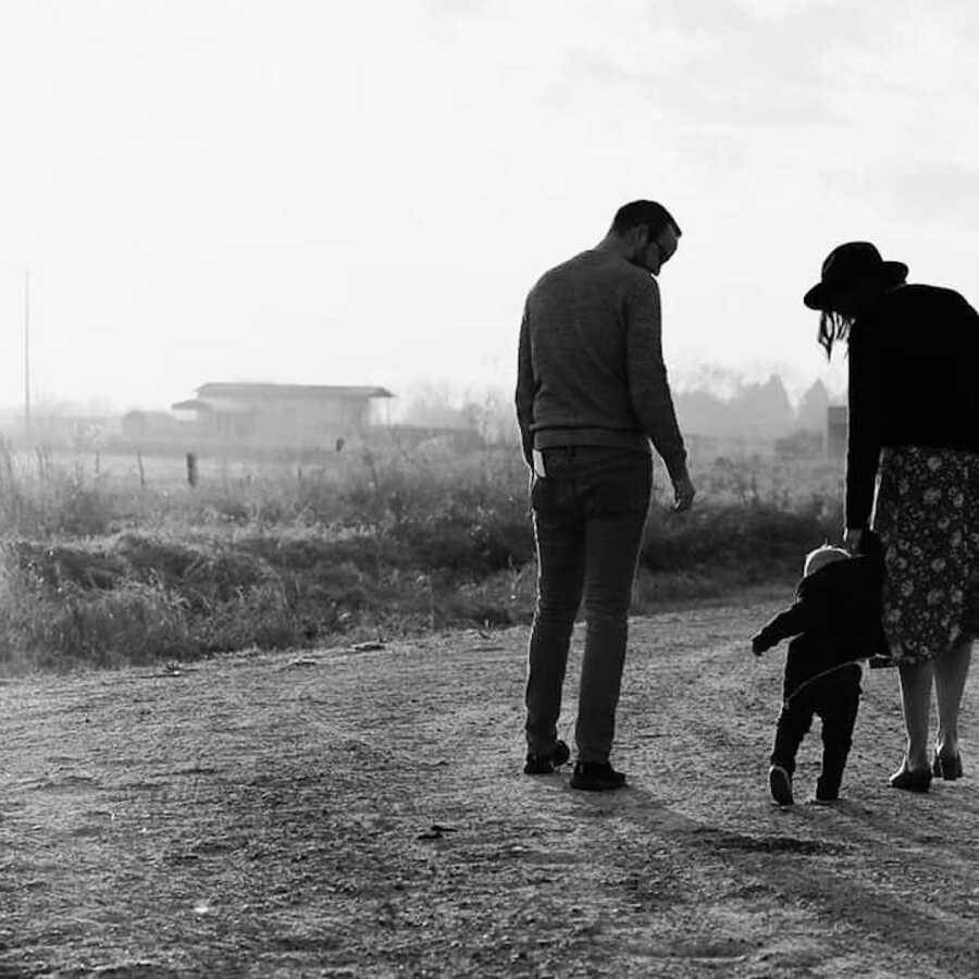 Husband and wife stand with their child, the mother is holding the child's hand