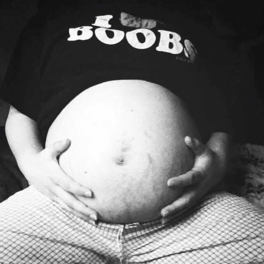 Pregnant woman cradles her pregnant stomach while wearing an "I heart boobs" t-shirt