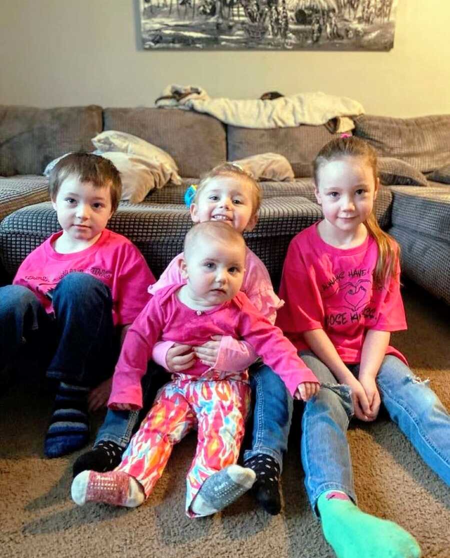 Four siblings sit on the floor and lean up against a brown couch while wearing matching pink shirts