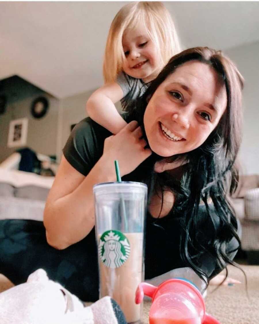 Mom crouches on the floor while her daughter climbs onto her back with a Starbucks drink in front of them