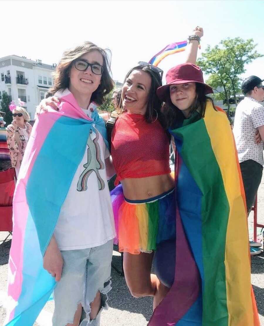 Mom in rainbow tutu takes a photo with her two daughters at a pride parade, one wrapped in the transgender pride flag and the other wrapped in the rainbow pride flag