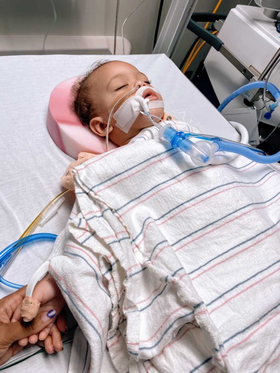 Young boy sleeps in the hospital while attached to wires and a breathing machine while his mom holds his hand