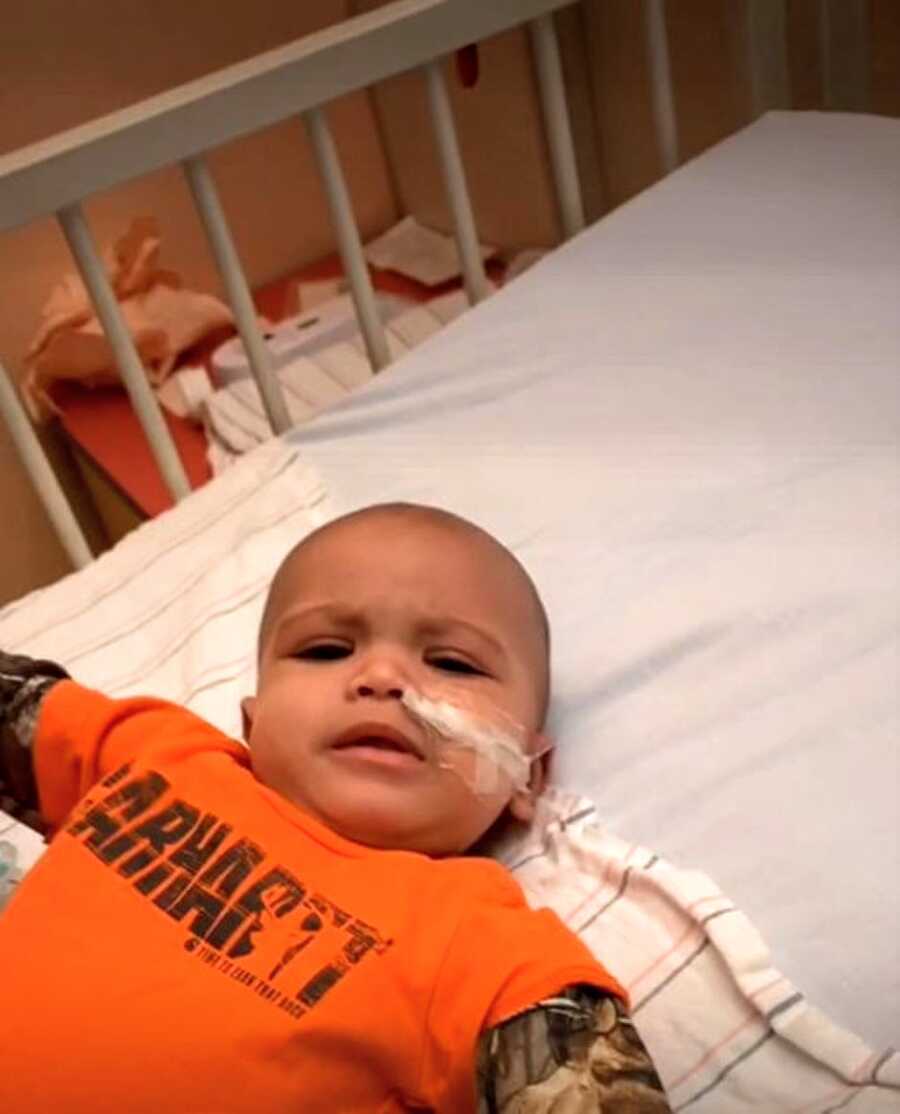 Young boy in bright orange Carhartt shirt looks grumpy while laying in a hospital crib