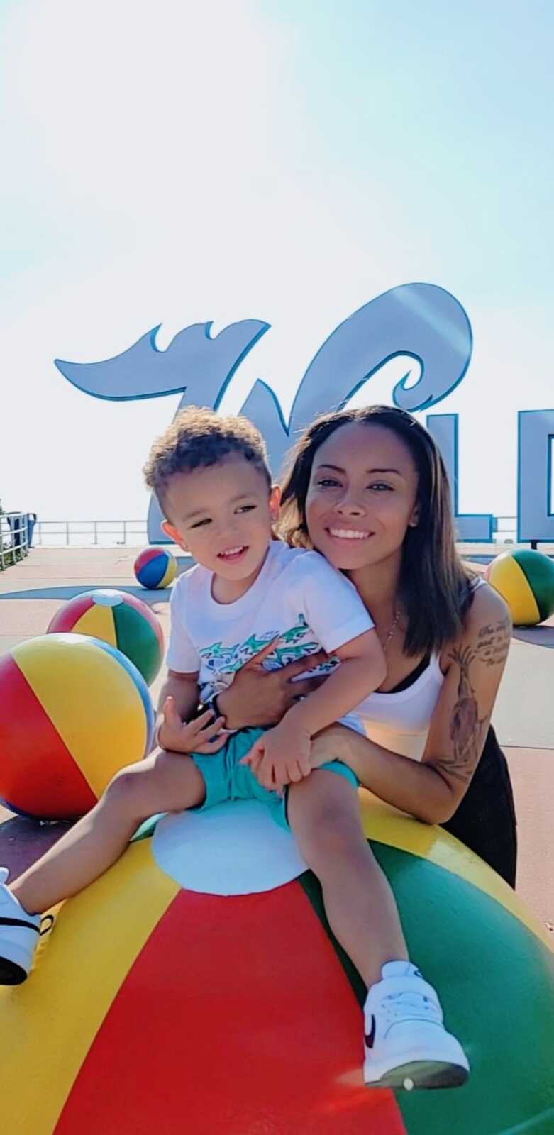 Mom and son take a photo together while on a beach vacation with beach balls and the ocean behind them