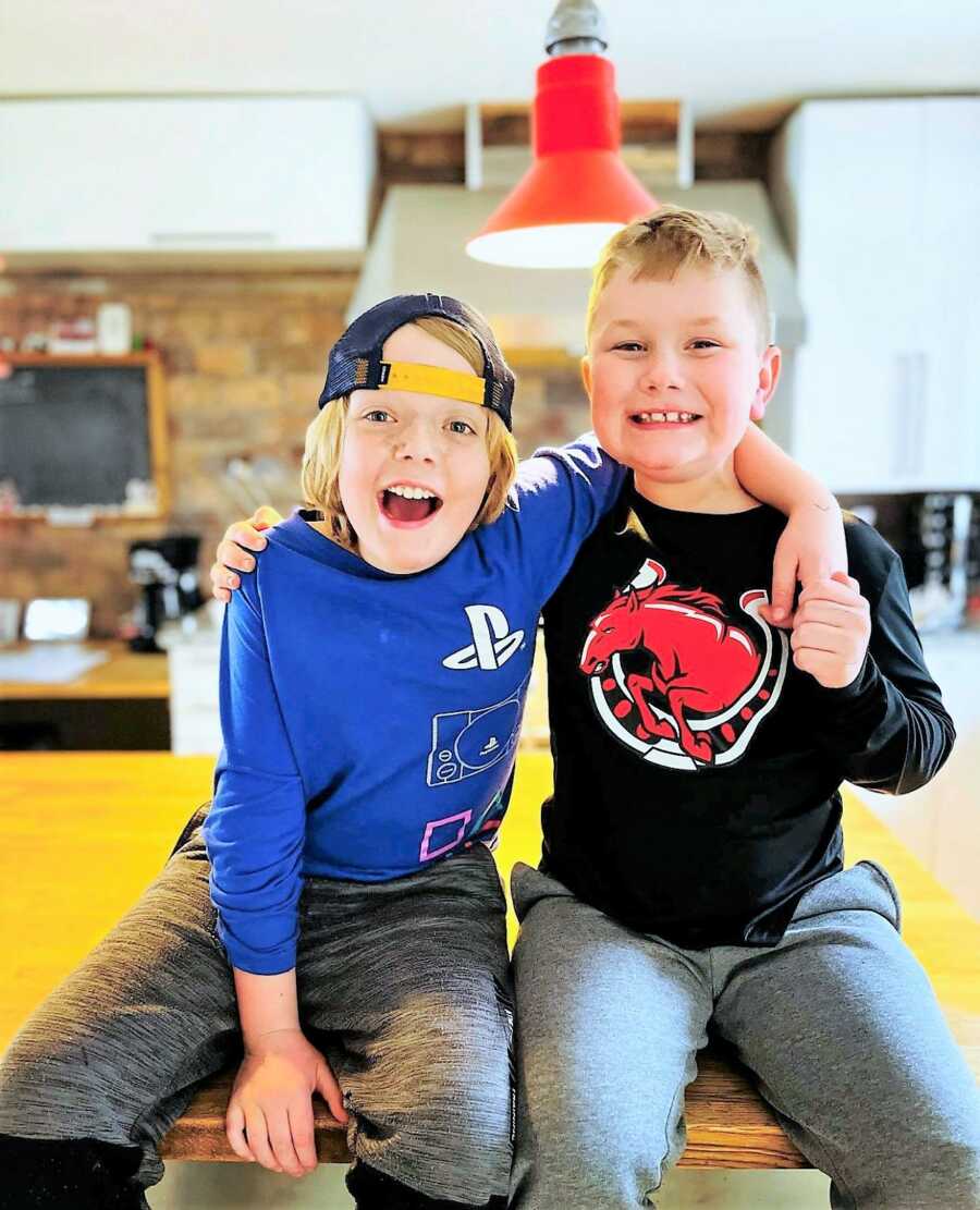 Two young brothers sitting on a kitchen countertop hugging each other and smiling big 