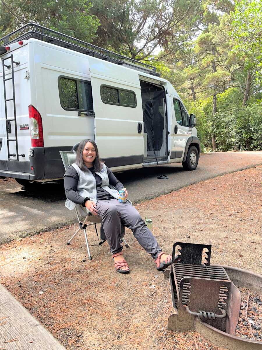 a girl sitting outside a white camper van in front of a small grilling spot