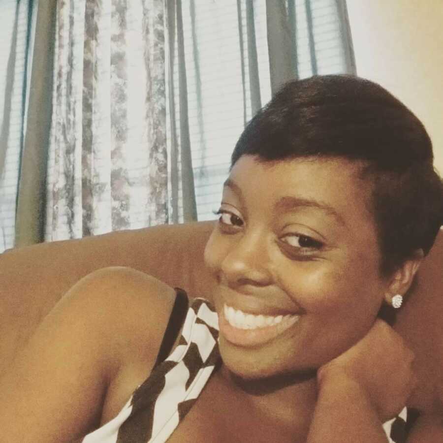 A young mom sits on a couch in front of grey curtains wearing a black and white tank top