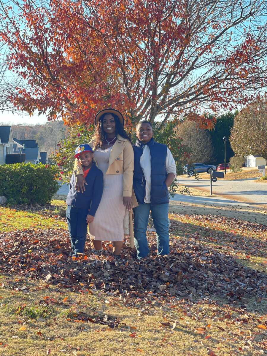 A mom stands in a leaf pile with her two sons in the fall