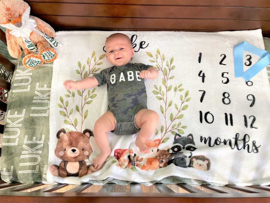 Baby boy wearing a camo print shirt with the word "BABE" and laying on a blanket printed with leaves, and wood animals