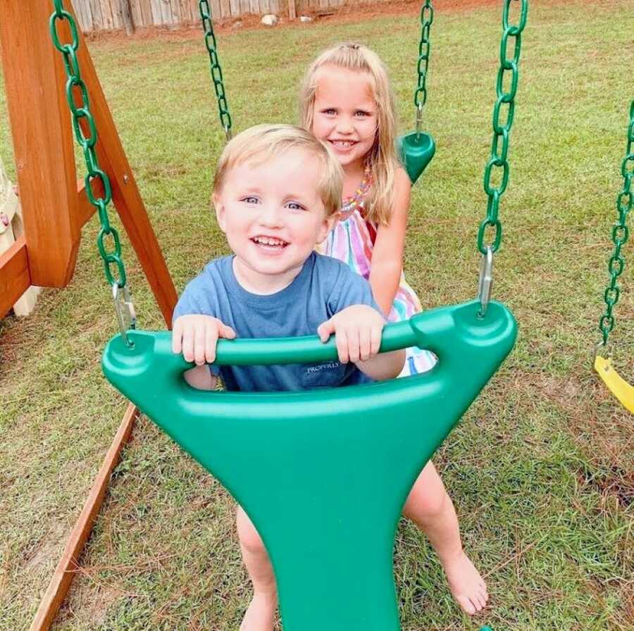 Brother and sister smile for a photo while on a green swing together in their backyard