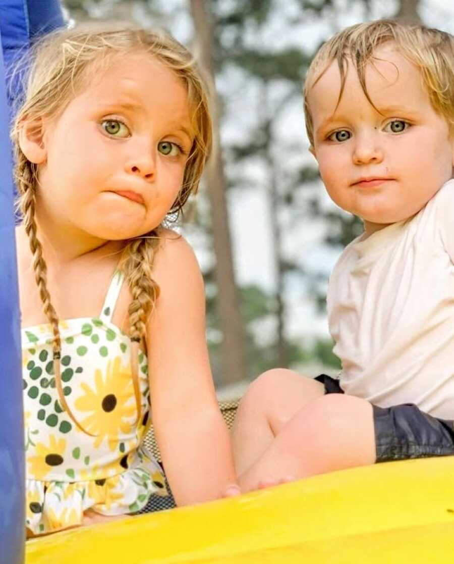 Big sister and little brother both stare into the camera for a photo while playing on a playground together