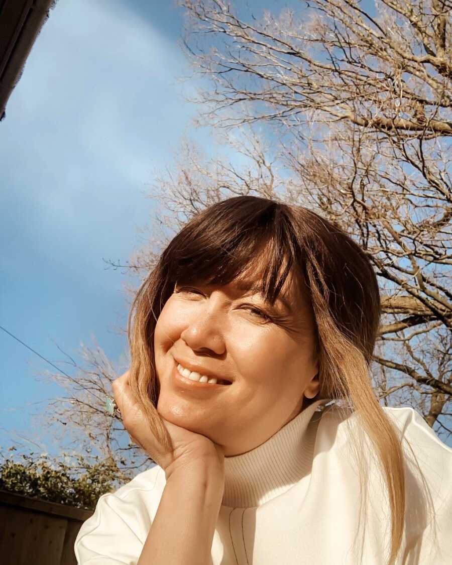 Woman battling incurable breast cancer smiles for a selfie while enjoying the sunshine in a white turtleneck sweater