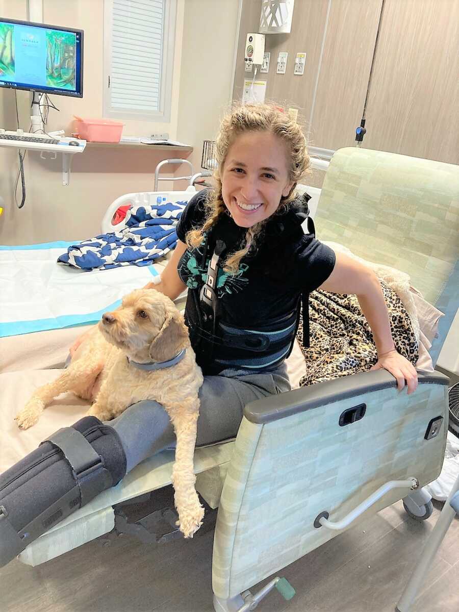 Young woman wearing a cast on her leg sitting in a hospital chair with her dog 