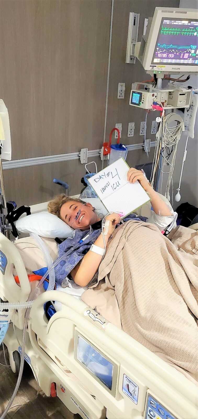 Young woman laying in a hospital bed holding a sing that says "DAY 4 ICU" and the date