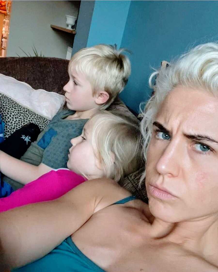 Mom of two looks angry in a selfie with her son and daughter in the background watching TV